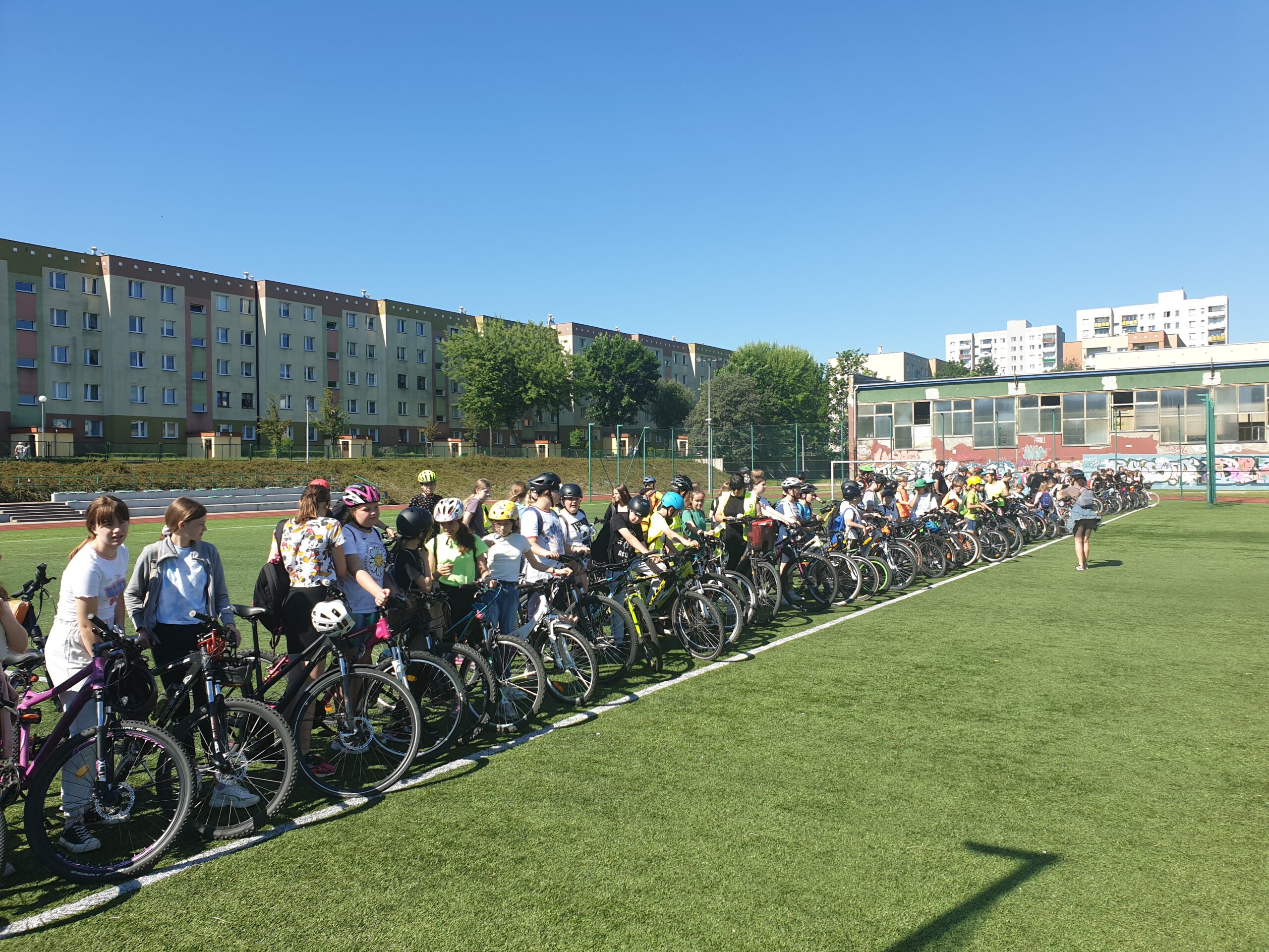Bardzo liczna grupa rowerzystów stoi ustawiona na płycie boiska