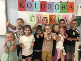 Grupa dziesięciu dzieci unosi kciuki do góry na tle napisu Kolorowa Chemia