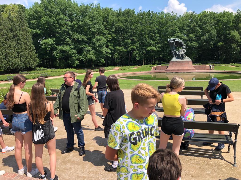 Uczestnicy wycieczki rozmawiają z przewodnikiem pod pomnikiem Chopina w Łazienkach w Warszawie