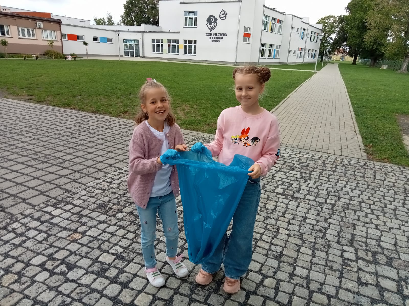 Dwie dziewczynki stoją z workiem na śmieci na tle budynku szkoły