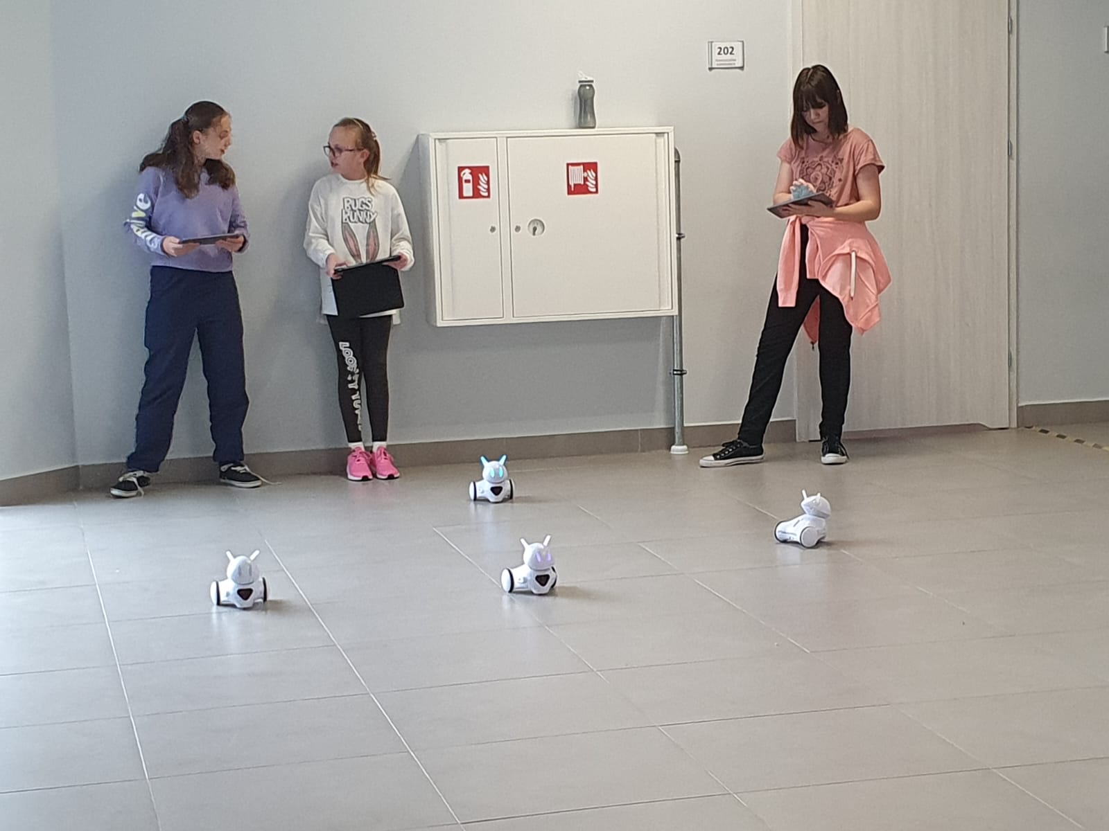 Trzy dziewczynki sterują swoimi robotami na szkolnym korytarzu